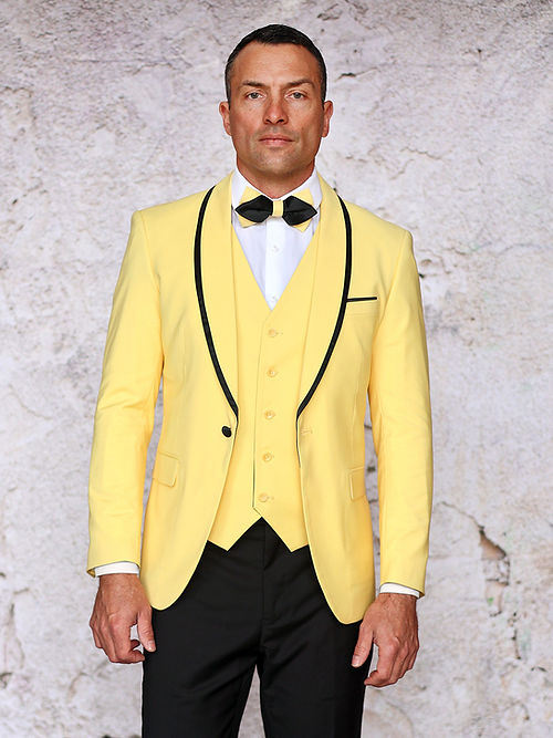 A man in a yellow Insomnia MANZINI SUNSET Slim Fit 3-Piece Tuxedo Suit with a black bow tie and black pants stands against a textured white backdrop, showcasing elegance with its reversible vest design.