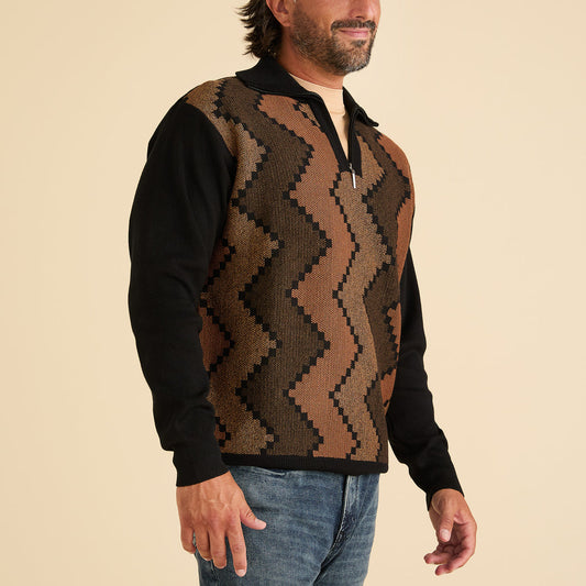 A man in an Inserch Zig-Zag Half Zip Sweater SW613-01 Black with a brown and black pattern on the front, paired with blue jeans, stands against a beige background.