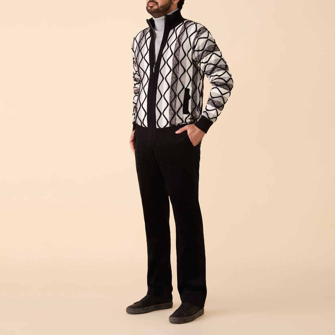 A man stands against a beige background wearing the Inserch Bouclé Yarn Graphic Full Zip Sweater (SW614-00001) in black, paired with a light gray turtleneck, black pants, and dark shoes.