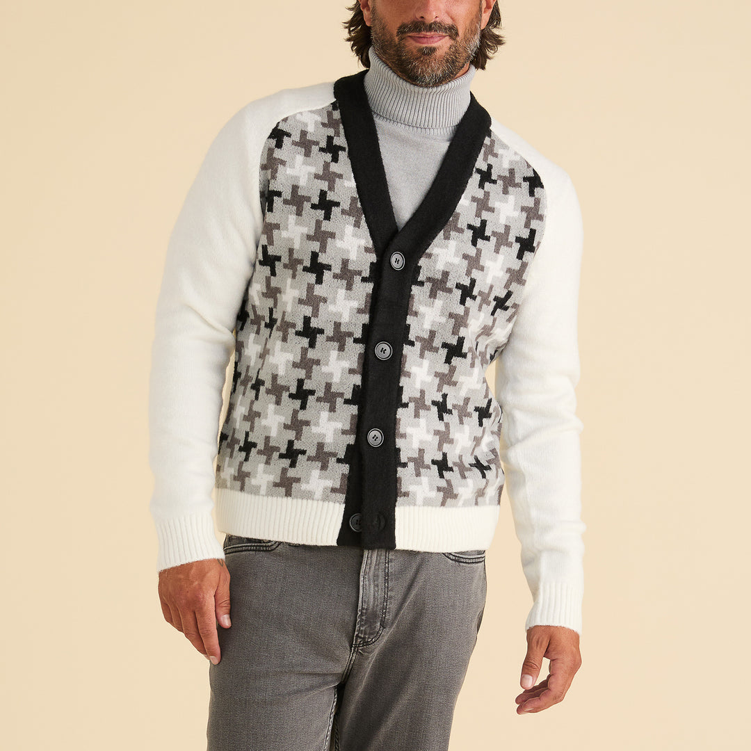 A man wears the Inserch Houndstooth Pattern Cardigan SW906-41 Black/White by Inserch, featuring black buttons and a black and gray cross pattern, styled with a light gray turtleneck and gray jeans against a beige background.