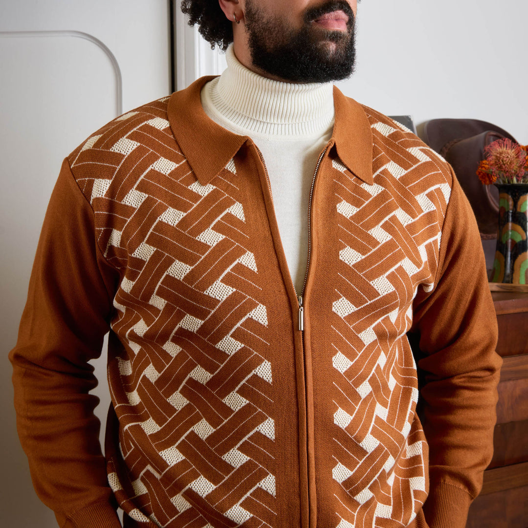 A man stands against a beige background in the Inserch Geometric Full Zip Sweater SW908-00005 Caramel, paired with a white turtleneck, light beige pants, and dark brown shoes. His face is partially out of frame.