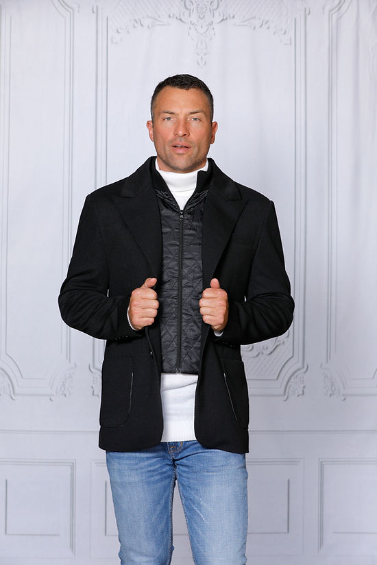 A man with short hair wears a MANZINI SWITZERLAND Black Blazer Coat with a zip-out liner from Insomnia, tailored fit vest, and jeans. He stands before an ornately decorated light gray wall, holding the coat's lapels.