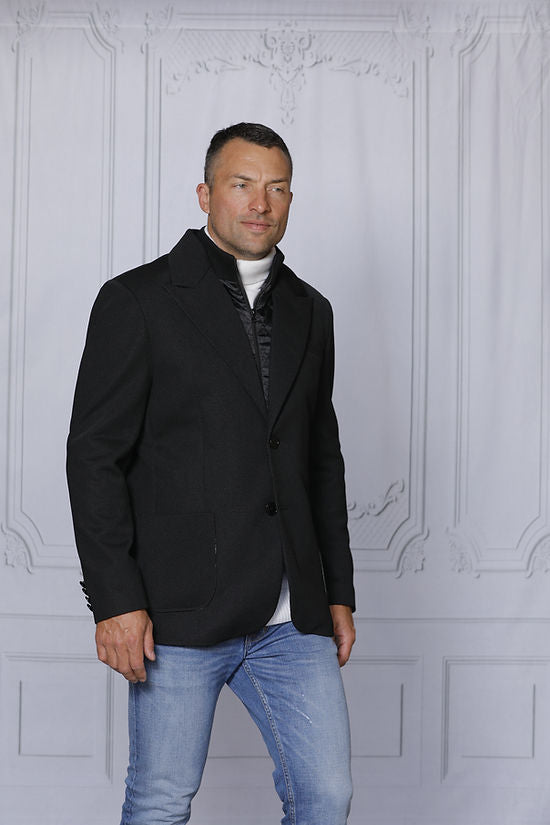 A man with short hair wears a MANZINI SWITZERLAND Black Blazer Coat with a zip-out liner from Insomnia, tailored fit vest, and jeans. He stands before an ornately decorated light gray wall, holding the coat's lapels.