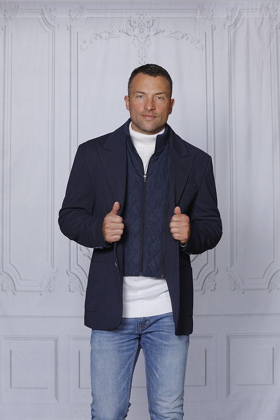 A man poses stylishly against a textured white wall, wearing the Insomnia MANZINI SWITZERLAND navy blazer coat with a zip-out liner over a white sweater.
