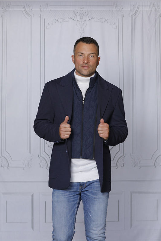 A man poses stylishly against a textured white wall, wearing the Insomnia MANZINI SWITZERLAND navy blazer coat with a zip-out liner over a white sweater.