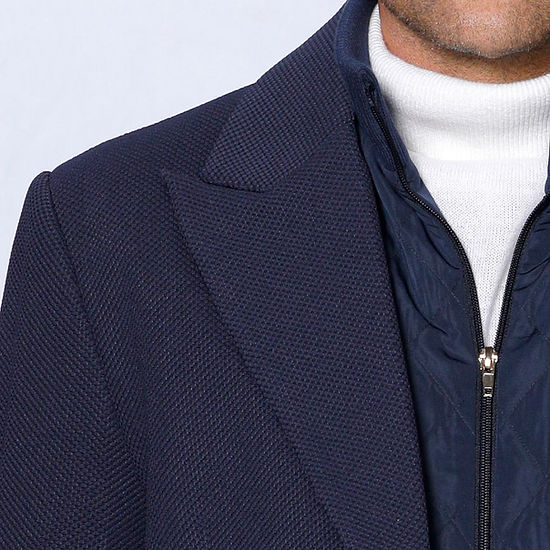 A man poses stylishly against a textured white wall, wearing the Insomnia MANZINI SWITZERLAND navy blazer coat with a zip-out liner over a white sweater.