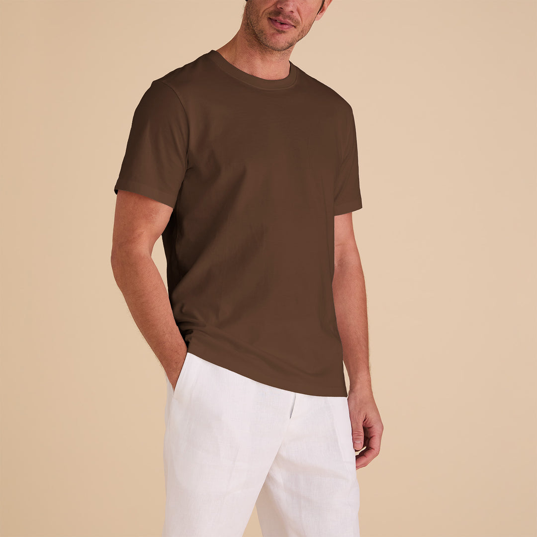 A man in a regular-fit Inserch T301-00064 Mocha T-shirt, made from plush Pima cotton, wears white pants against a beige background, casually tucking his left hand into his pocket.