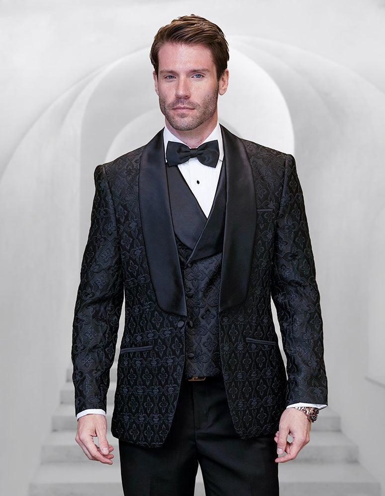 A man wearing the STATEMENT CLOTHING | TIZANO-BLACK tuxedo by Statement Clothing, featuring black lace, a satin shawl lapel, and a black bow tie, stands before a white arched background.
