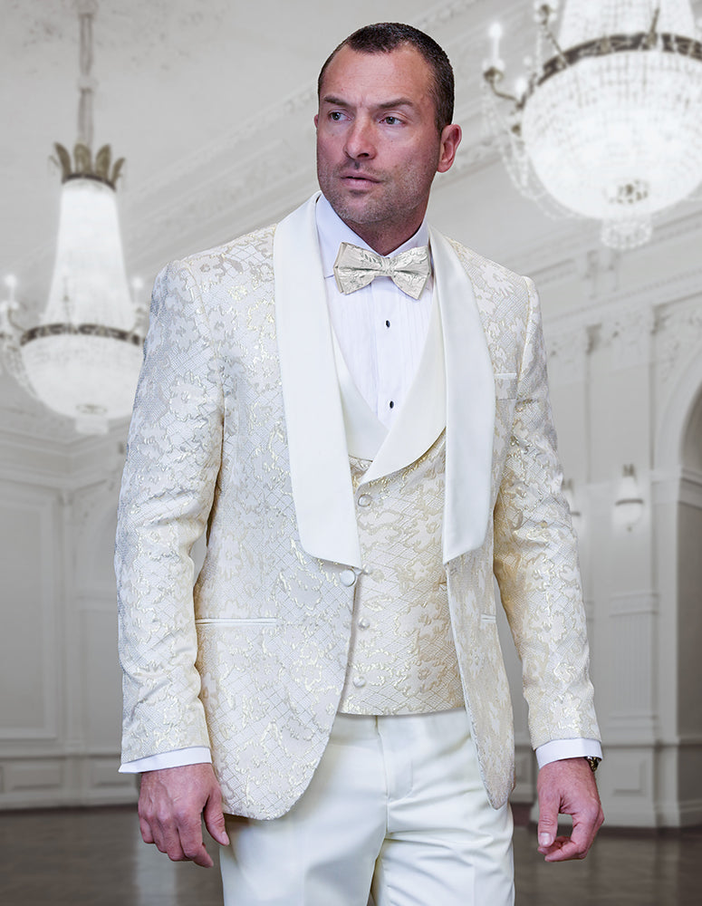 A man in the STATEMENT CLOTHING | TIZANO-OFFWHITE modern fit tuxedo by Statement Clothing, with a bow tie, stands in an elegant, chandelier-lit ballroom.