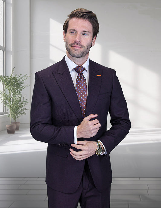 A man in a modern fit, peak lapel STATEMENT CLOTHING | TRIESTE-EGGPLANT suit by Statement Clothing stands indoors before light walls and windows, adjusting his sleeve. A patterned tie and a potted plant appear in the background.