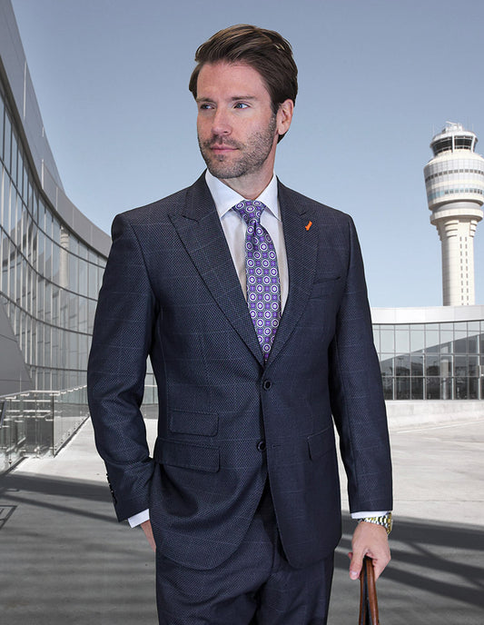 A man in the Statement Clothing TRIESTE-GRAY suit, paired with a patterned tie, stands near a modern glass building and air traffic control tower, holding an eggplant-colored item in his left hand.