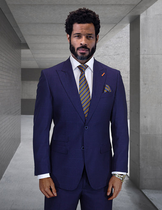 A man with short curly hair and a beard models the STATEMENT CLOTHING | TRIESTE-NAVY suit by Statement Clothing, paired with a white shirt and patterned tie, while standing in a modern concrete hallway.