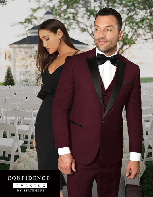 A man wears the STATEMENT CLOTHING | TUX-PK-BURGUNDY, a maroon tuxedo with black lapels, as he stands before a woman in black at an outdoor wedding. Text: "CONFIDENCE EVENING BY STATEMENT." Tailored fit for timeless elegance.