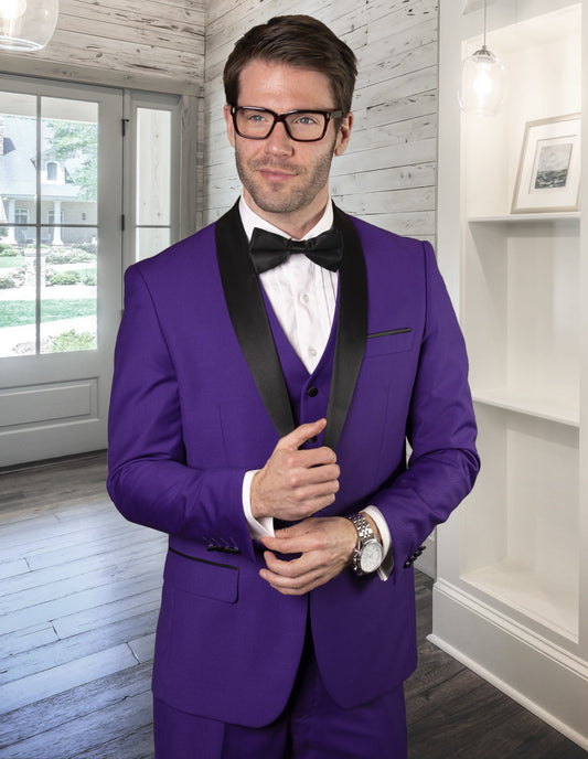 A man wearing the STATEMENT CLOTHING | TUX-SH-PURPLE by Statement Clothing with black shawl lapels, a white shirt, black bow tie, and glasses stands indoors adjusting his jacket.