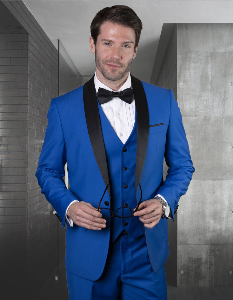 A man indoors, holding eyeglasses, wears the STATEMENT CLOTHING | TUX-SH-ROYALBLUE by Statement Clothing—a bright blue tailored fit tuxedo with a black shawl lapel and bow tie.