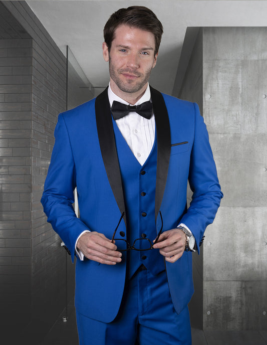 A man indoors, holding eyeglasses, wears the STATEMENT CLOTHING | TUX-SH-ROYALBLUE by Statement Clothing—a bright blue tailored fit tuxedo with a black shawl lapel and bow tie.