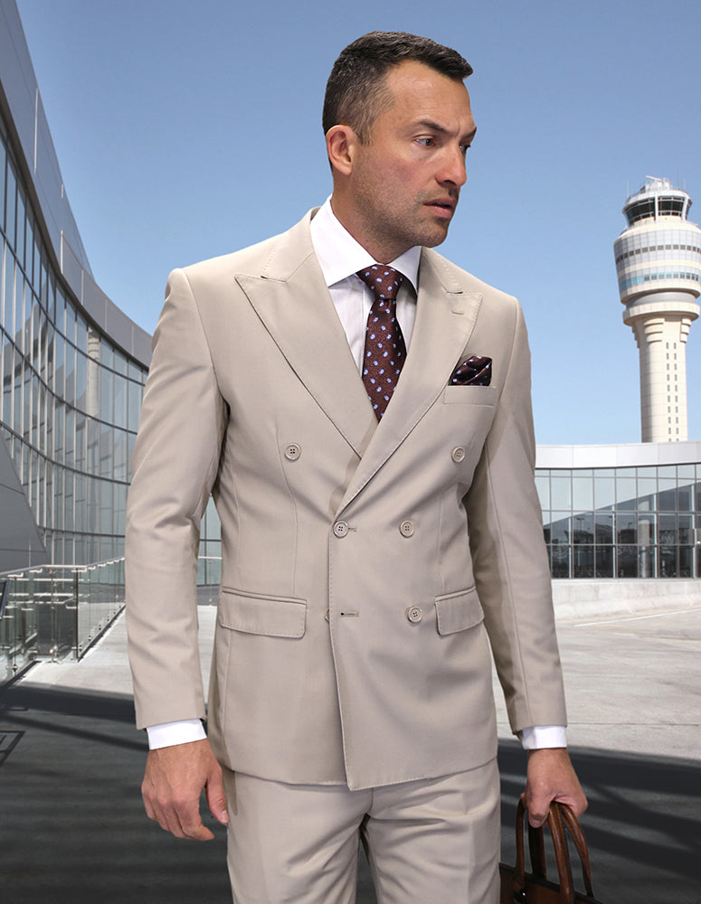 A man wears the STATEMENT CLOTHING | TZD-100-TAN modern fit, double-breasted light beige suit by Statement Clothing, holding a brown bag outside a glass building with an airport control tower in the background.