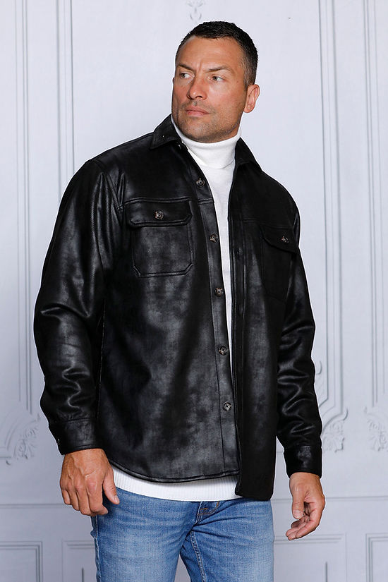 A man in a black vegan suede spandex sweater shirt by Insomnia and a white turtleneck stands against a white paneled wall, looking to his right. He is wearing blue jeans.