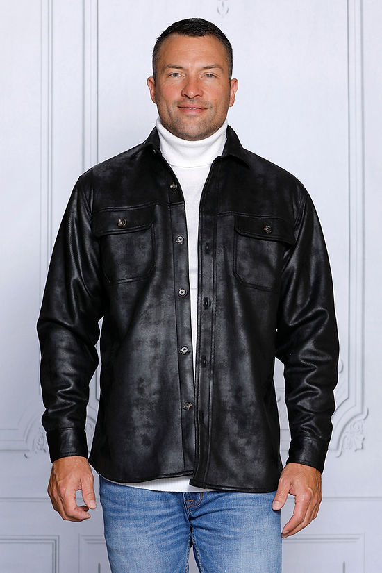 A man in a black vegan suede spandex sweater shirt by Insomnia and a white turtleneck stands against a white paneled wall, looking to his right. He is wearing blue jeans.