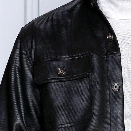 A man in a black vegan suede spandex sweater shirt by Insomnia and a white turtleneck stands against a white paneled wall, looking to his right. He is wearing blue jeans.