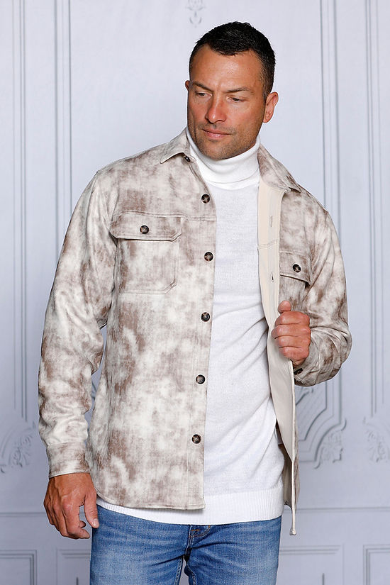 A man in a MANZINI VALENTINO Tan Soft Vegan Suede Button Sweater Shirt by Insomnia, paired with blue jeans, stands against a textured white wall.