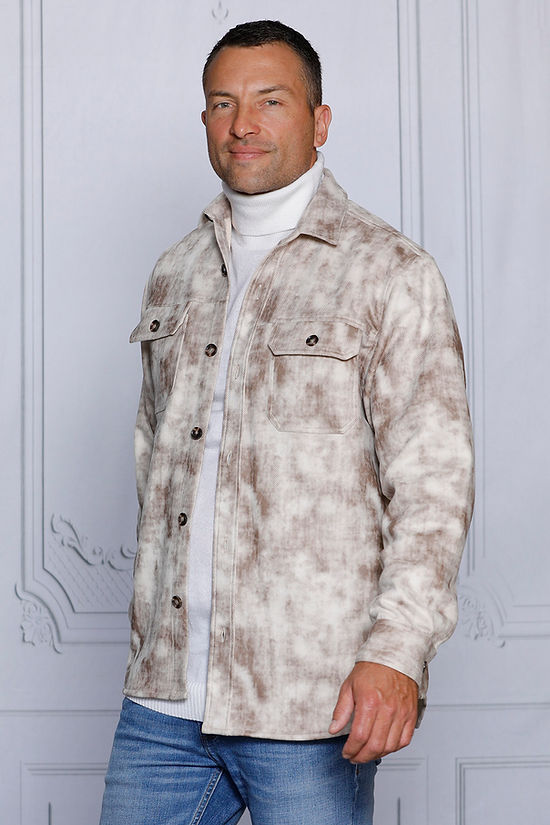 A man in a MANZINI VALENTINO Tan Soft Vegan Suede Button Sweater Shirt by Insomnia, paired with blue jeans, stands against a textured white wall.