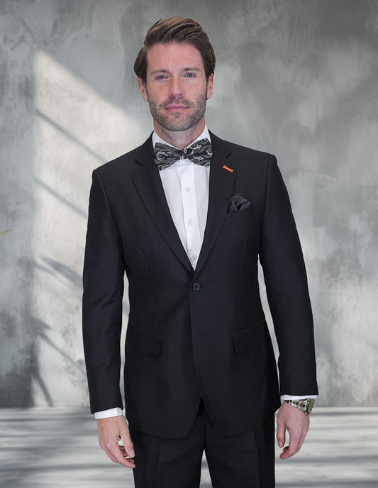 A man wears the STATEMENT CLOTHING | VERNAZA-BLACK modern fit suit by Statement Clothing, tailored from Italian Super 200's Wool, with a white shirt and patterned bow tie, standing indoors against a gray textured background.