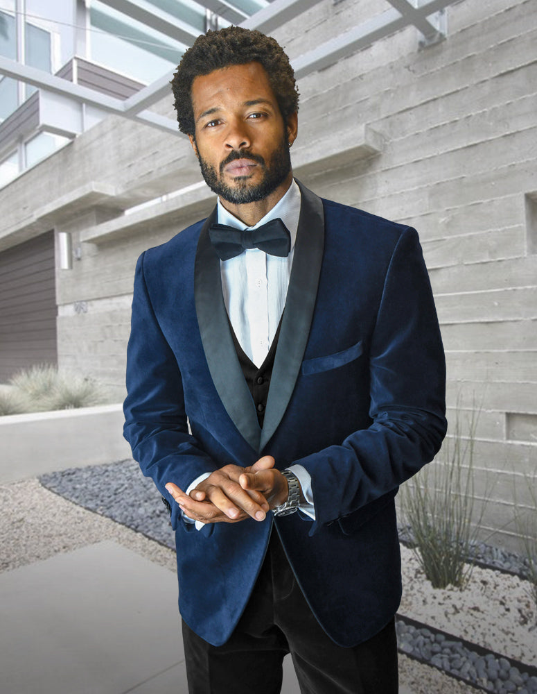 A man wearing the Statement Clothing VL-100-NAVY tailored fit velvet suit jacket and bow tie stands outdoors before a modern building with concrete walls and landscaping.
