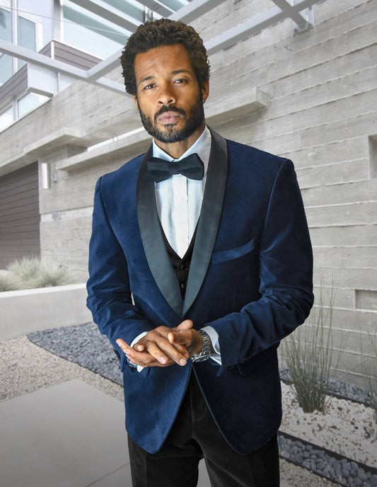 A man wearing the Statement Clothing VL-100-NAVY tailored fit velvet suit jacket and bow tie stands outdoors before a modern building with concrete walls and landscaping.