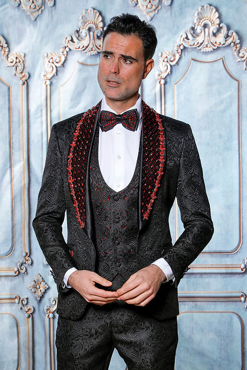 A man poses against ornate moldings in an Insomnia MANZINI ZARA 3-piece slim fit tuxedo, featuring a black/red floral pattern with rhinestone trim, and a bow tie.