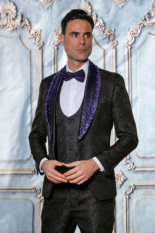 A man in an Insomnia MANZINI ZARA 3-piece slim fit rhinestone floral pattern black/royal tuxedo suit with a blue polka dot bow tie stands against an ornate backdrop.