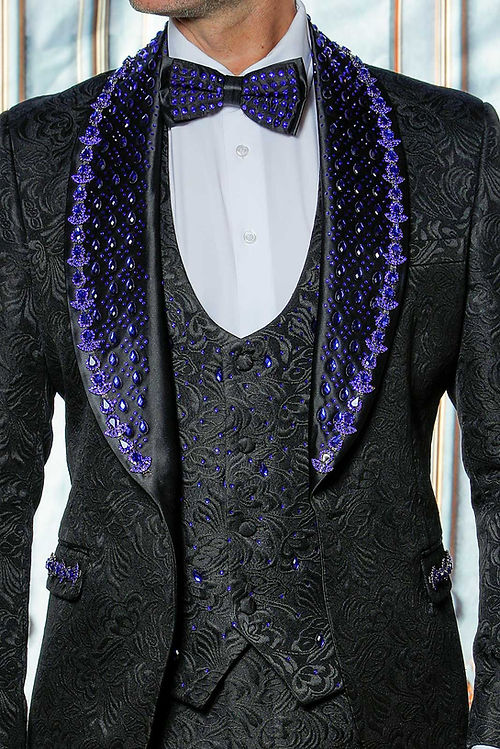 A man in an Insomnia MANZINI ZARA 3-piece slim fit rhinestone floral pattern black/royal tuxedo suit with a blue polka dot bow tie stands against an ornate backdrop.