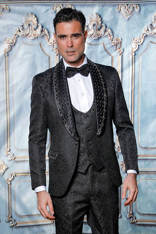 A man wearing an Insomnia MANZINI ZARA 3-Piece Slim Fit Rhinestone Floral Pattern Black Tuxedo Suit and a bow tie stands before a decorative backdrop.