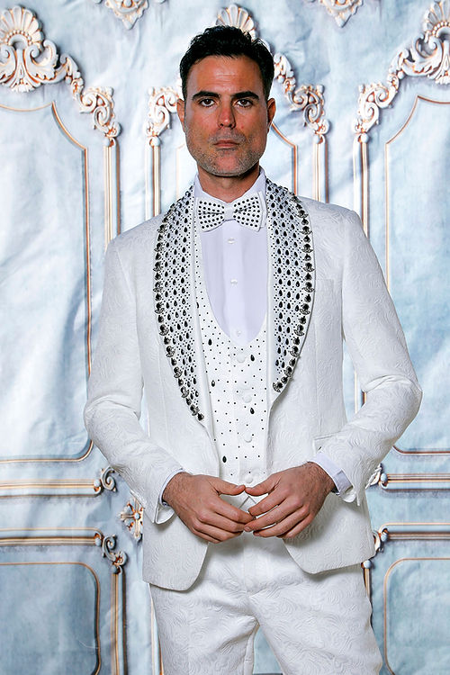 A man in an Insomnia MANZINI ZARA 3-piece slim fit white tuxedo with rhinestone floral pattern and black accents, paired with a polka dot bow tie, stands against an intricate background.