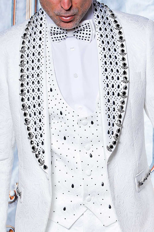 A man in an Insomnia MANZINI ZARA 3-piece slim fit white tuxedo with rhinestone floral pattern and black accents, paired with a polka dot bow tie, stands against an intricate background.