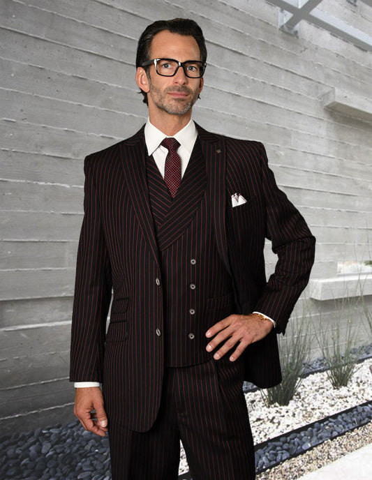 A man wearing the Statement Clothing ZARELLI-B-RED dark pinstripe suit with regular fit pleated pants, white shirt, and maroon tie stands before a modern concrete wall amid gravel and sparse plants.