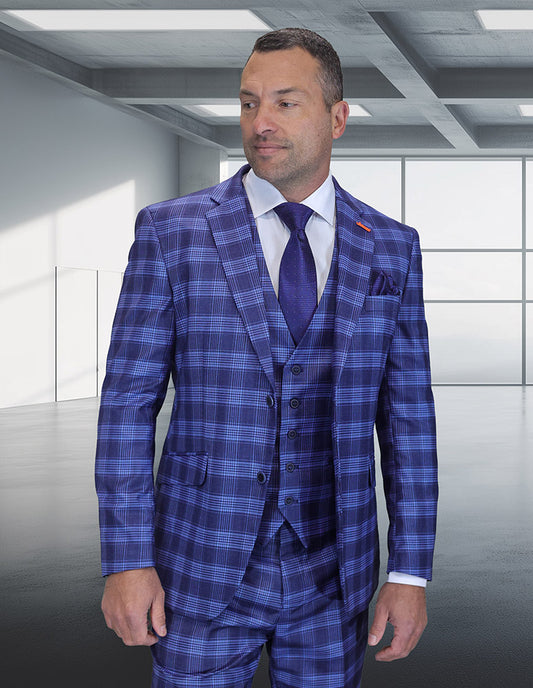 A man in the STATEMENT CLOTHING | ZILO-1-INDIGO modern fit suit by Statement Clothing, paired with a white shirt and blue tie, stands in a sleek room with large windows and a concrete ceiling.