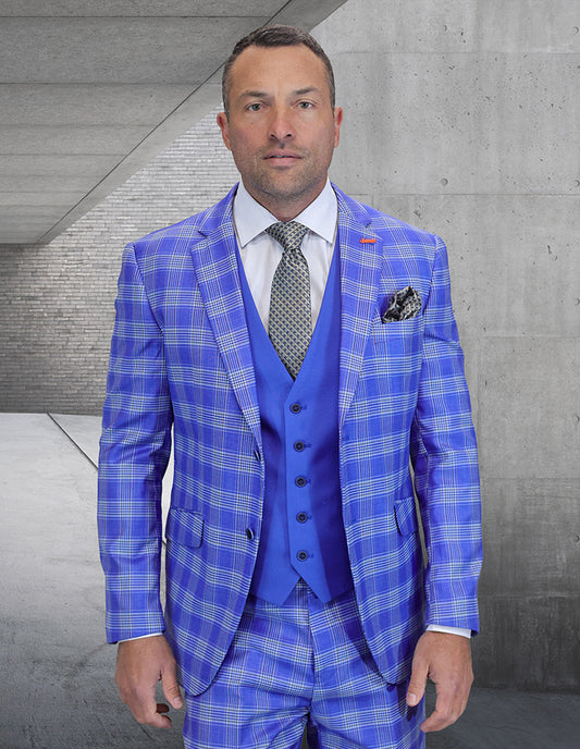 A man wearing the STATEMENT CLOTHING | ZILONI-2-STEELBLUE modern fit Italian wool suit by Statement Clothing, featuring a bright blue plaid jacket, blue vest, white shirt, and patterned tie, stands against a concrete and brick backdrop.