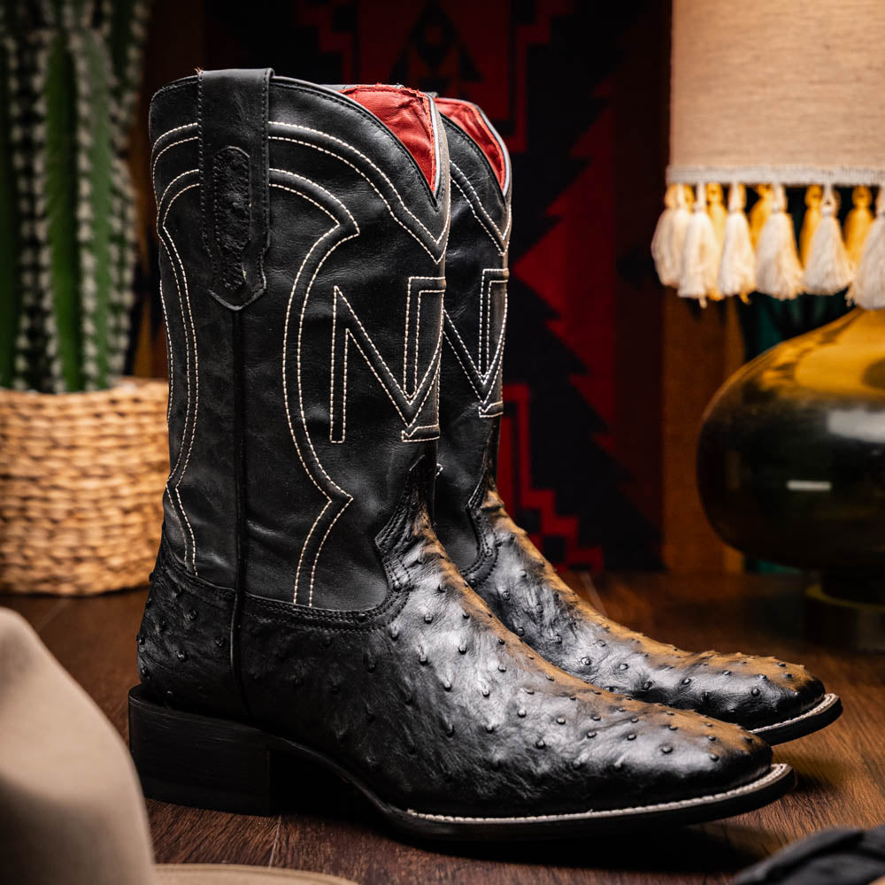 The Alabama Black by Marco Di Milano: a pair of black ostrich skin cowboy boots with white stitching, shown from side and sole views. These luxury men's boots combine classic western style with superb craftsmanship.