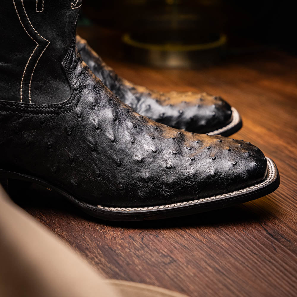 The Alabama Black by Marco Di Milano: a pair of black ostrich skin cowboy boots with white stitching, shown from side and sole views. These luxury men's boots combine classic western style with superb craftsmanship.