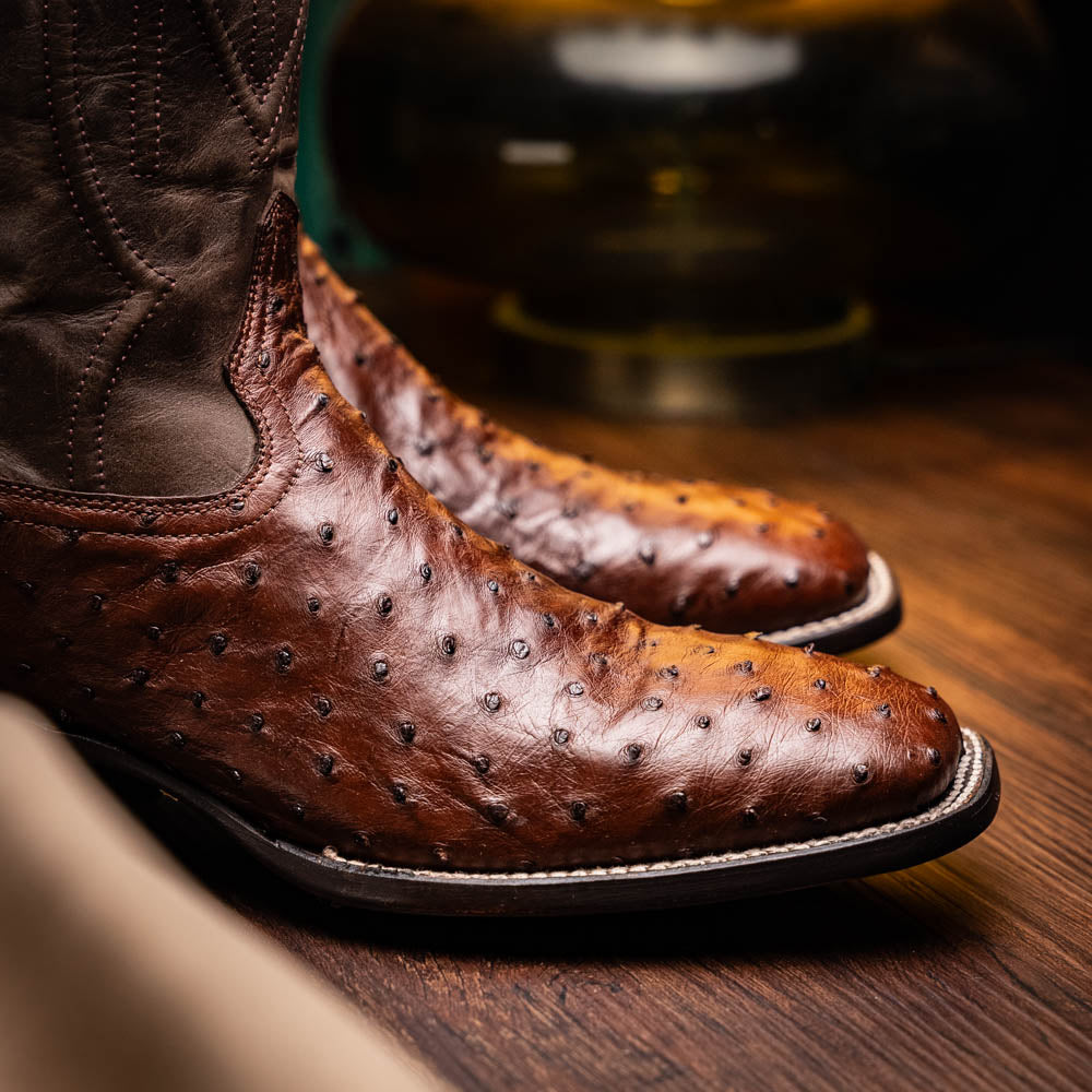 A pair of Marco Di Milano Alabama Kango Tabac men’s boots, handcrafted with genuine ostrich skin foot and dark upper, one upright and one displaying the branded sole, showcase classic Western cowboy style.