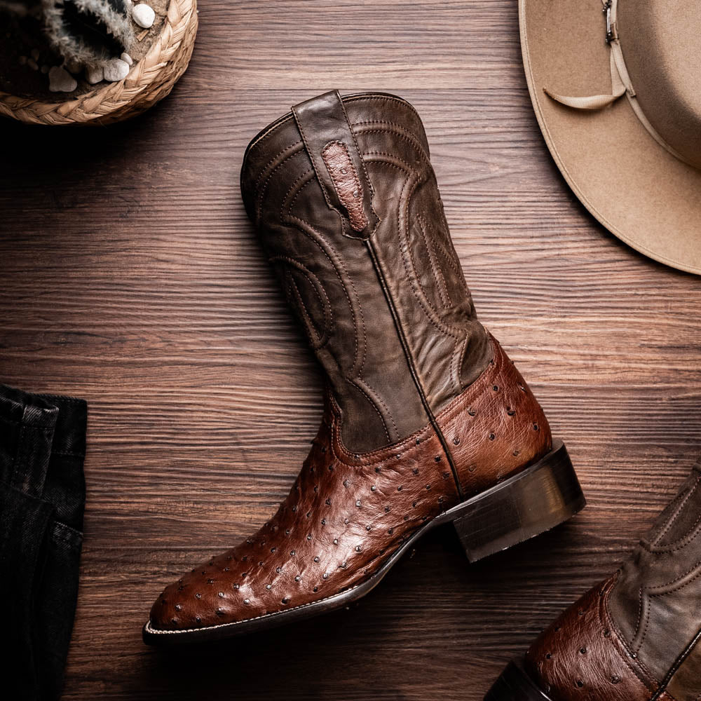 A pair of Marco Di Milano Alabama Kango Tabac men’s boots, handcrafted with genuine ostrich skin foot and dark upper, one upright and one displaying the branded sole, showcase classic Western cowboy style.