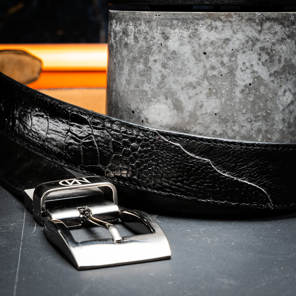 Alessio Belt Black by Marco Di Milano—a handcrafted leather belt in black with a textured finish and a silver buckle engraved with “ATELIER MILANO.” The belt is artfully coiled in a loose loop against a white background.