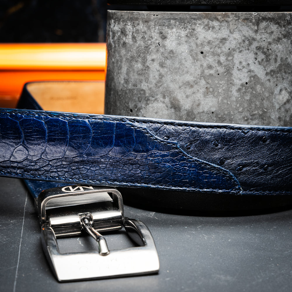 The Alessio Belt Navy by Marco Di Milano is a coiled blue handcrafted luxury belt with a silver buckle, featuring both smooth and textured crocodile-patterned sections.
