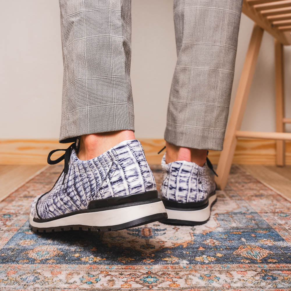 The Alonzo Newspaper sneakers by Marco Di Milano feature genuine caiman crocodile in white and black with a hand-painted finish, black laces, and black rubber soles, displayed with one shoe propped on the other against a white background.