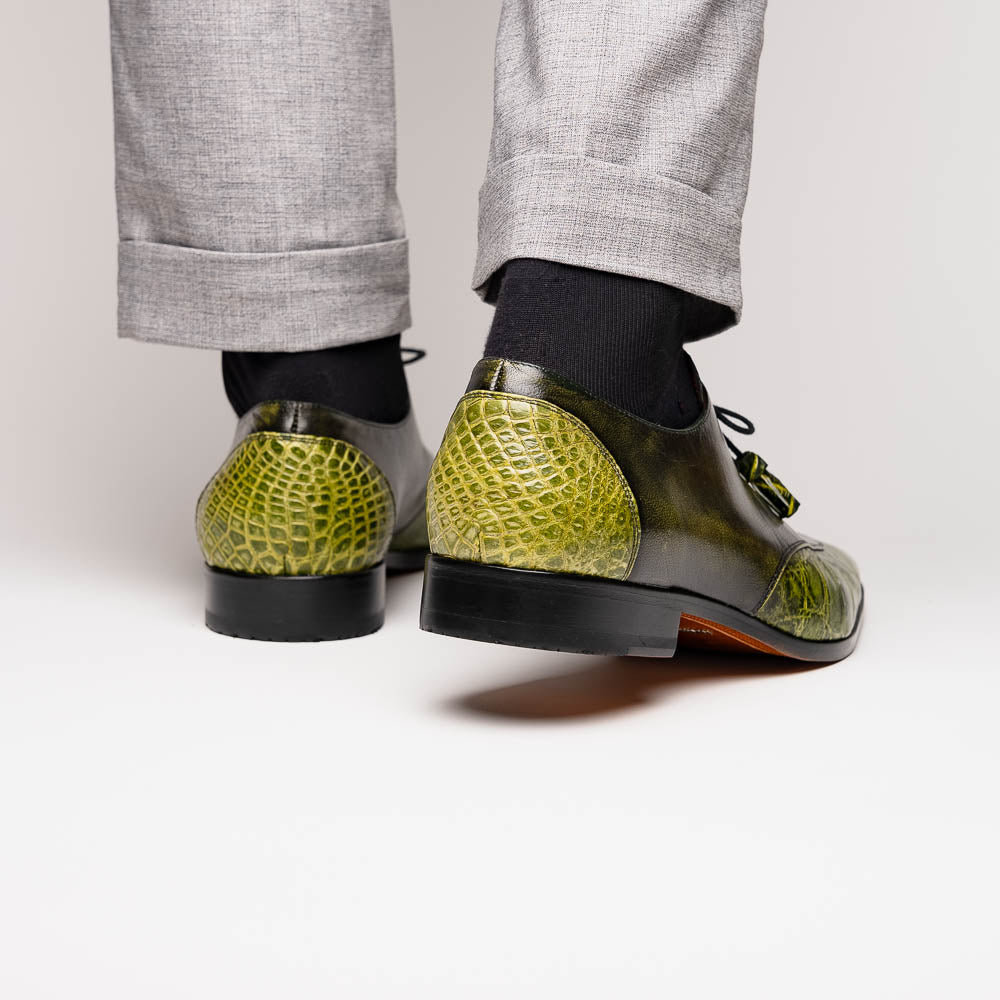 A pair of Anzio Rustic Green men's dress shoes by Marco Di Milano, featuring a green textured finish, black laces, and brown leather soles—one upright and one lying on its side.