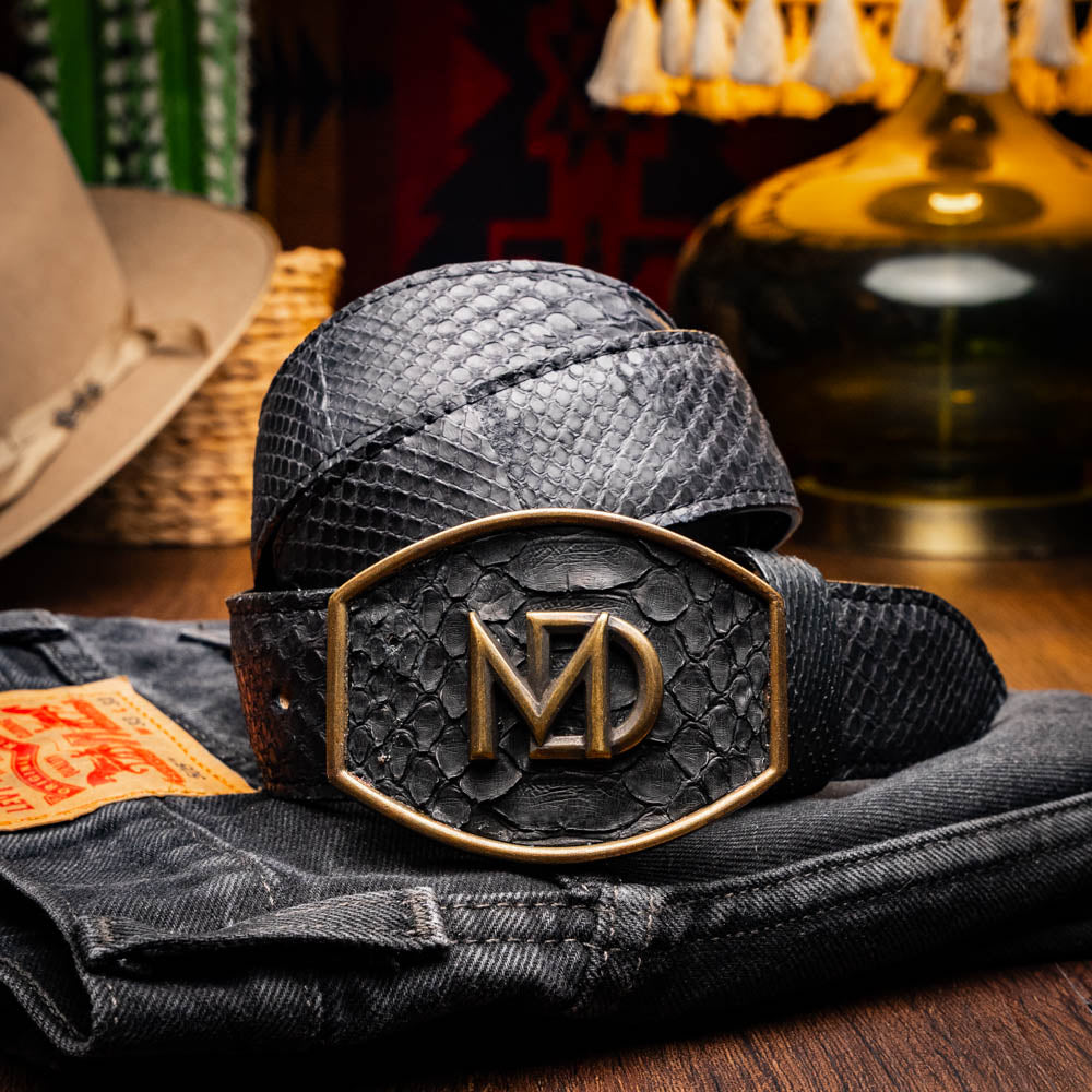 The Arkansas Belt Black by Marco Di Milano features textured black leather and a gold MD monogram buckle, shown coiled on a white background.