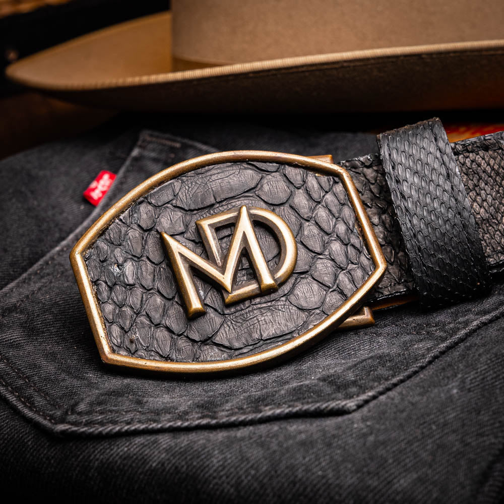 The Arkansas Belt Black by Marco Di Milano features textured black leather and a gold MD monogram buckle, shown coiled on a white background.