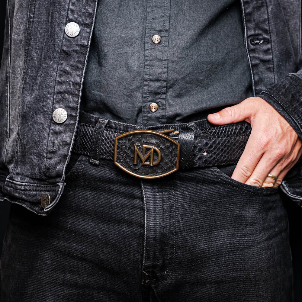 The Arkansas Belt Black by Marco Di Milano features textured black leather and a gold MD monogram buckle, shown coiled on a white background.