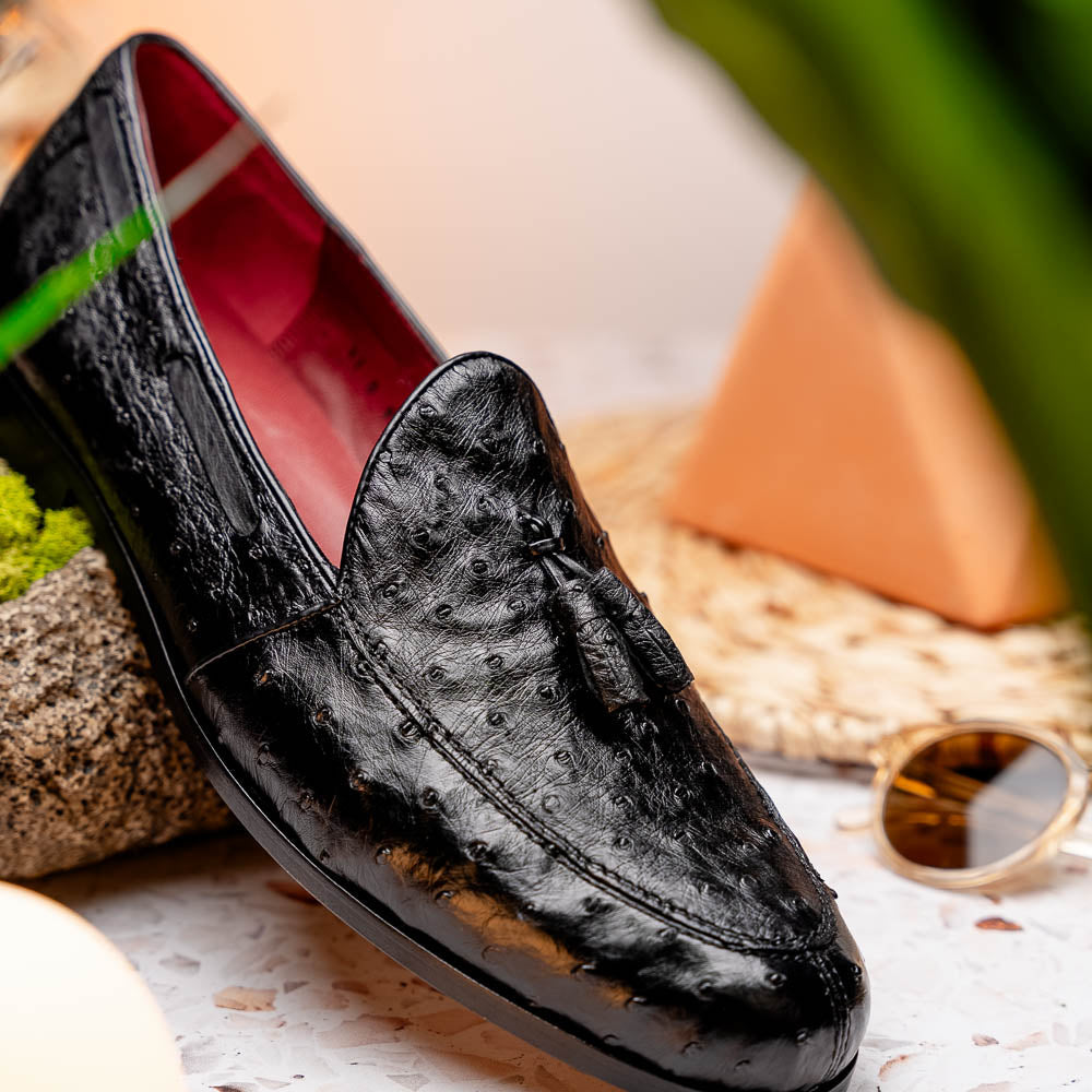 The Aubiere Black by Marco Di Milano are luxury men's tassel loafers in black genuine ostrich leather, shown with one shoe upright and the other displaying a tan sole embossed with the Marco Di Milano logo.